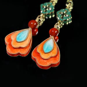 May include: Close-up of a pair of ornate earrings. Each earring features a layered, petal-shaped design in shades of orange and a central turquoise accent. Red beads and gold-colored metal complete the design.