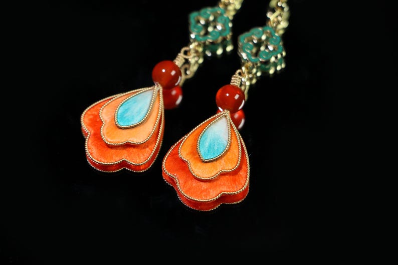 May include: Close-up of a pair of ornate earrings. Each earring features a layered, petal-shaped design in shades of orange and a central turquoise accent. Red beads and gold-colored metal complete the design.
