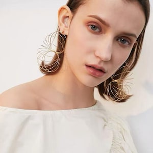 May include: A pair of gold-toned earrings featuring a multi-layered, spiralling wire design that creates a circular, cage-like structure. The earrings are constructed from thin, metallic wire loops that overlap to form a lightweight, open-work sphere.