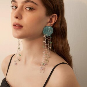 Op de afbeelding: Hangende oorbellen met een blauwgroene, geschulpte schelpvormige bovenkant versierd met kleine witte sterretjes. Meerdere delicate zilverkleurige kettinkjes hangen aan de schelp, verfraaid met kleine, doorschijnende, groenachtige edelsteensplinters en kralen.