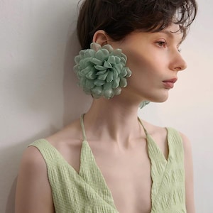 May include: A large, sage green floral earring made of layered, translucent fabric petals. The earring features a dense, rounded bloom design with a soft, matte texture. It is worn on the earlobe, showcasing a voluminous, multi-layered construction.