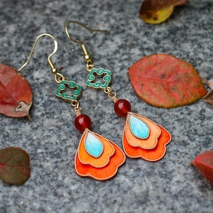 May include: A pair of gold-toned dangle earrings with orange, turquoise, and red accents. The earrings feature a layered petal design with a turquoise teardrop shape at the center. The earrings are set against a gray stone surface with fallen leaves.
