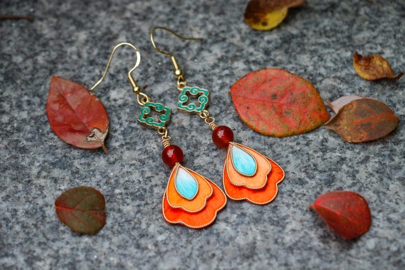May include: A pair of gold-toned dangle earrings with orange, turquoise, and red accents. The earrings feature a layered petal design with a turquoise teardrop shape at the center. The earrings are set against a gray stone surface with fallen leaves.