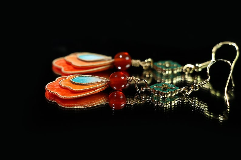 May include: A pair of gold-toned earrings with a layered, fan-shaped design in orange, with a turquoise teardrop accent. The earrings feature red beads and turquoise square accents.