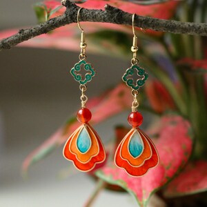 May include: A pair of gold-toned dangle earrings. Each earring features a teal and orange fan-shaped pendant, a red bead, and a green filigree accent. The earrings are suspended from gold-colored hooks.