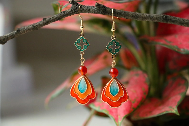 May include: A pair of gold-toned dangle earrings. Each earring features a teal and orange fan-shaped pendant, a red bead, and a green filigree accent. The earrings are suspended from gold-colored hooks.