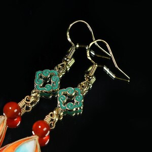May include: Gold-toned dangle earrings with intricate details. The earrings feature turquoise and orange enamel accents, red beads, and decorative square elements with turquoise enamel. The design includes a chain and hook closure.