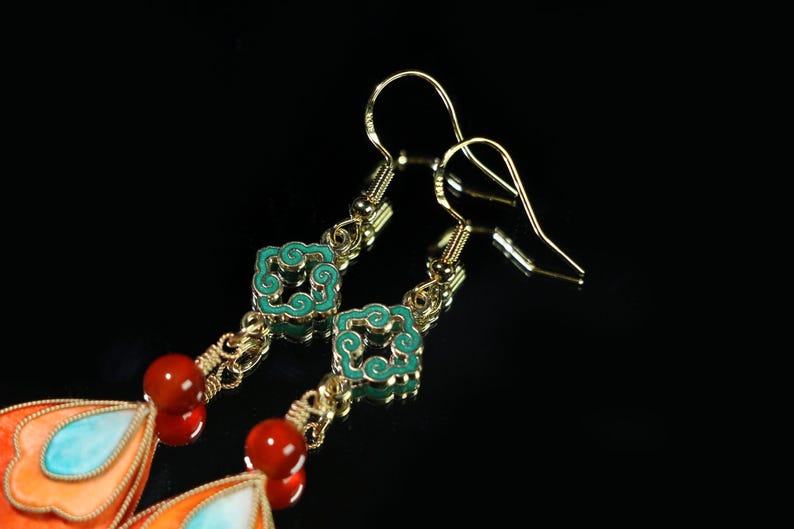May include: Gold-toned dangle earrings with intricate details. The earrings feature turquoise and orange enamel accents, red beads, and decorative square elements with turquoise enamel. The design includes a chain and hook closure.