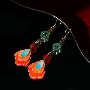 May include: A pair of dangle earrings with a gold-tone finish. Each earring features a layered, orange and blue pendant, a red bead, and a turquoise floral accent. The earrings are set against a black background with a red accent.
