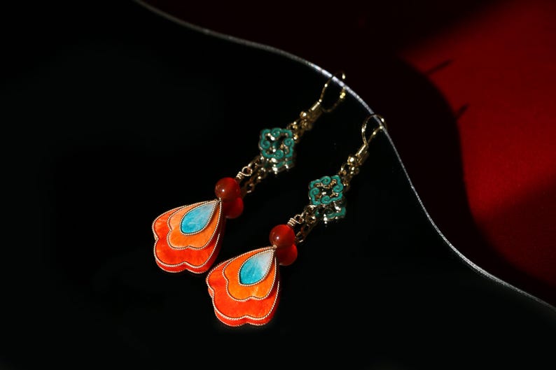 May include: A pair of dangle earrings with a gold-tone finish. Each earring features a layered, orange and blue pendant, a red bead, and a turquoise floral accent. The earrings are set against a black background with a red accent.
