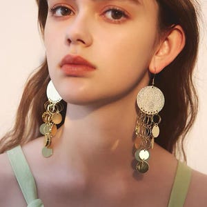 May include: A pair of gold-toned dangle earrings featuring a large circular top disc connected to multiple smaller, dangling circular discs that create a layered, fringe-like effect. The metal surfaces have a reflective, polished finish.