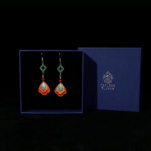 May include: A pair of colorful earrings with a unique design, displayed in a blue box. The earrings feature orange, turquoise, and red accents, with a decorative green cross-shaped element. The box has the text "Toscane Blanc" on it.