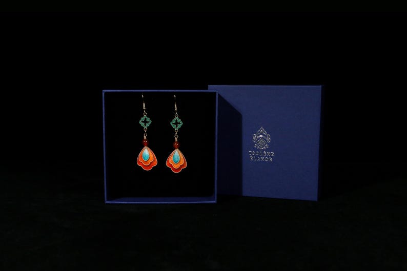 May include: A pair of colorful earrings with a unique design, displayed in a blue box. The earrings feature orange, turquoise, and red accents, with a decorative green cross-shaped element. The box has the text "Toscane Blanc" on it.