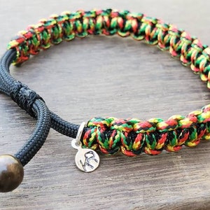 May include: A black paracord bracelet with red, yellow, and green woven into the design. The bracelet has wooden beads on each end and a silver charm with the letters "KR" on it.
