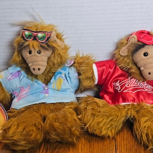 May include: Two plush ALF dolls are displayed. One wears a blue Hawaiian shirt and sunglasses, the other a red jacket with white trim and the text "Kidbutters". Both have brown fur and tan snouts.