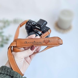 Könnte beinhalten: Ein brauner Leder-Kameragurt mit weißen Nähten und dem Text "Christina H Photography" sowie einem Herzsymbol. Der Gurt ist an einer schwarzen Sony-Kamera befestigt.