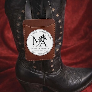 Könnte beinhalten: Ein brauner Lederkartenhalter mit Logo ist an einem schwarzen Cowboy-Stiefel mit Stern- und Blumendesigns befestigt. Der Kartenhalter hat ein rundes Logo mit der Aufschrift "Leather Works" und "Your Custom Design".