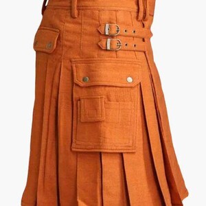 May include: Orange kilt with pleats, pockets, and buckle details. The kilt features a structured waistband with belt loops and two decorative buckles. The front has two pockets with button closures. This garment is a traditional style.