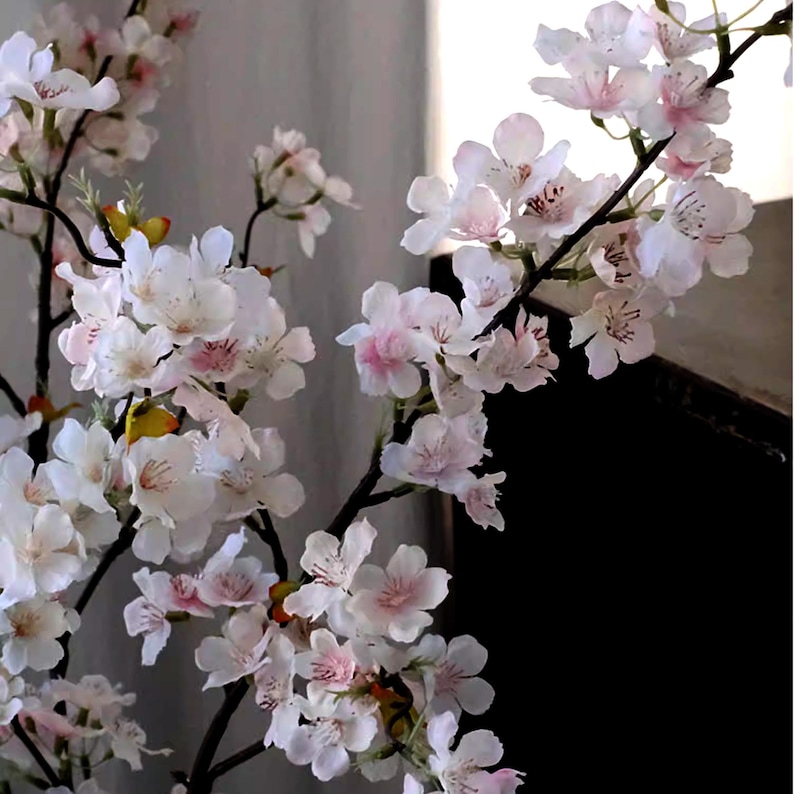May include: Close-up of artificial cherry blossom branches with white and pink flowers. The flowers have delicate petals and are clustered along the dark brown branches. The background is a neutral gray.