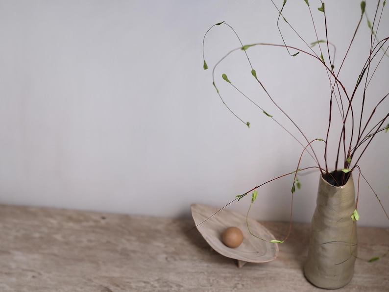 May include: A still life arrangement featuring a beige ceramic vase holding slender, arching branches with small green buds. A wooden, leaf-shaped dish holds a brown egg. The objects are set on a light-colored wooden surface.