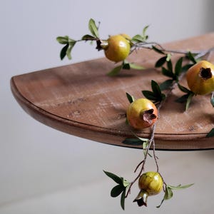 May include: A close-up of a wooden table with a branch of pomegranates. The pomegranates are yellow and orange with green leaves. The table is brown and has a rounded edge.