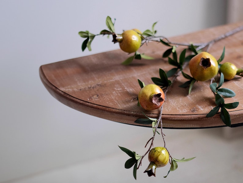 May include: A close-up of a wooden table with a branch of pomegranates. The pomegranates are yellow and orange with green leaves. The table is brown and has a rounded edge.