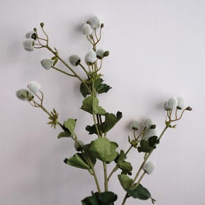 May include: A sprig of artificial flowers with green leaves and stems. The flower heads are small, round, and covered in a soft, fuzzy texture. The leaves are a deep green color. The arrangement is set against a plain white background.