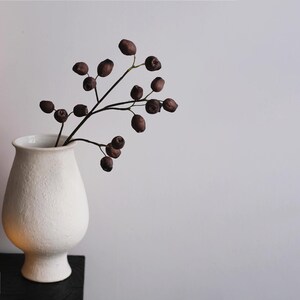 May include: A white ceramic vase with a textured surface holds a branch with dark brown berries. A lit candle sits on a black surface in the foreground. The background is a plain white wall.