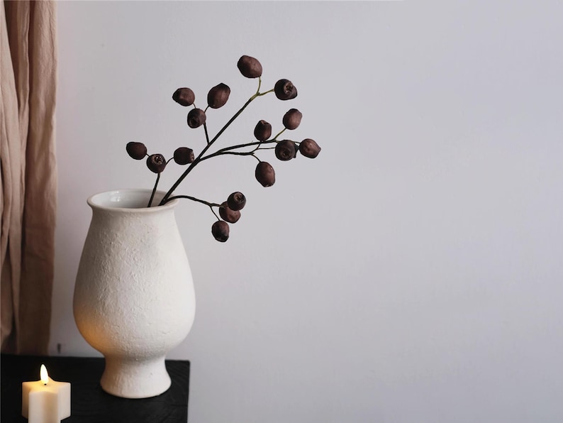 May include: A white ceramic vase with a textured surface holds a branch with dark brown berries. A lit candle sits on a black surface in the foreground. The background is a plain white wall.
