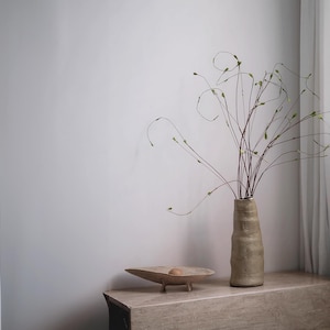May include: A beige ceramic vase with a textured surface holds slender branches with small green buds. A wooden bowl with a small sphere sits on a stone-like surface. The scene is set against a neutral wall and a white curtain.