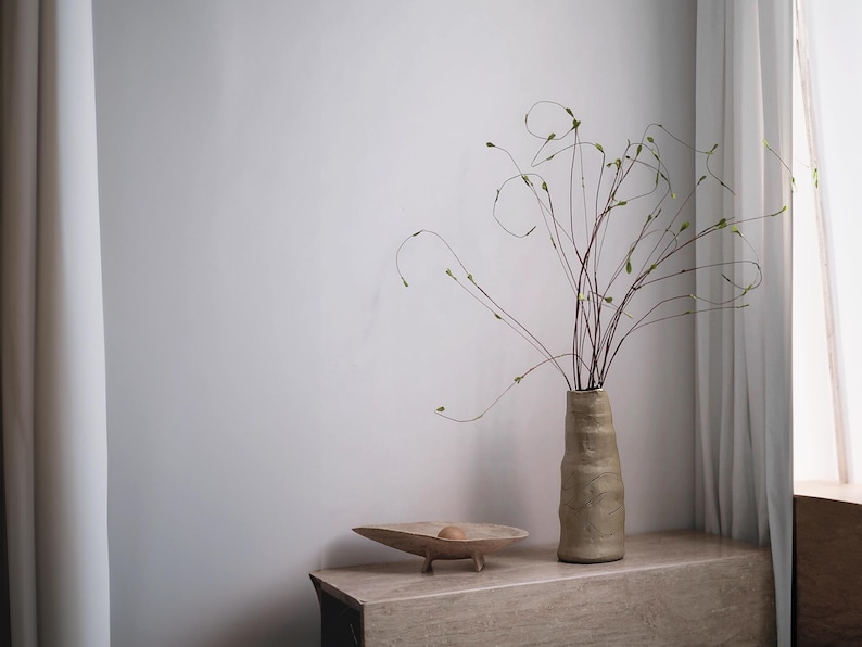 May include: A beige ceramic vase with a textured surface holds slender branches with small green buds. A wooden bowl with a small sphere sits on a stone-like surface. The scene is set against a neutral wall and a white curtain.