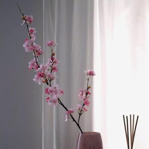 May include: A decorative arrangement featuring a branch of pink cherry blossoms in a mauve vase. The blossoms are in full bloom, with delicate petals and a natural appearance. The background includes a white curtain and a hint of a reed diffuser.