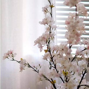 May include: Close-up of artificial cherry blossom branches with delicate white and pink flowers. The branches have dark stems and are set against a blurred background of a white curtain and a window with blinds.