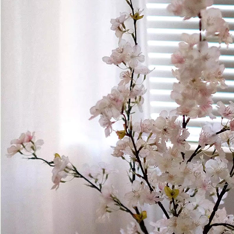 May include: Close-up of artificial cherry blossom branches with delicate white and pink flowers. The branches have dark stems and are set against a blurred background of a white curtain and a window with blinds.