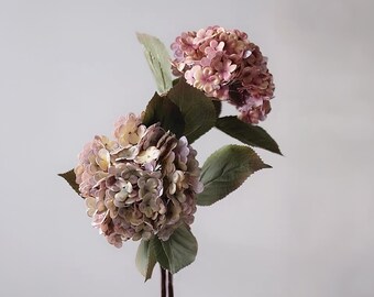May include: Two faux hydrangea flower stems with large, round blooms in shades of pink, lavender, and cream. The flowers have multiple layers of petals and are surrounded by large, green leaves. The stems are a dark brown color.