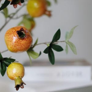 May include: Close-up of a decorative pomegranate branch with green leaves and several pomegranates. The pomegranates are orange and yellow with a speckled texture. The branch is a home decor item.
