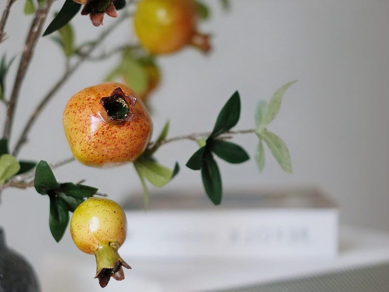 May include: Close-up of a decorative pomegranate branch with green leaves and several pomegranates. The pomegranates are orange and yellow with a speckled texture. The branch is a home decor item.