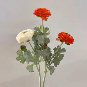 May include: Artificial ranunculus stems with three vibrant orange and one cream-coloured flower heads. The faux flowers have green leaves and buds, creating a realistic floral arrangement. Ideal for home decor or crafting.
