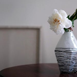 Pode incluir: Um vaso decorativo branco e preto com uma flor de pe&oacute;nia branca e um bot&atilde;o floral. O vaso tem um acabamento brilhante com um design linear preto na metade inferior. O vaso est&aacute; sobre uma mesa de madeira castanha escura.