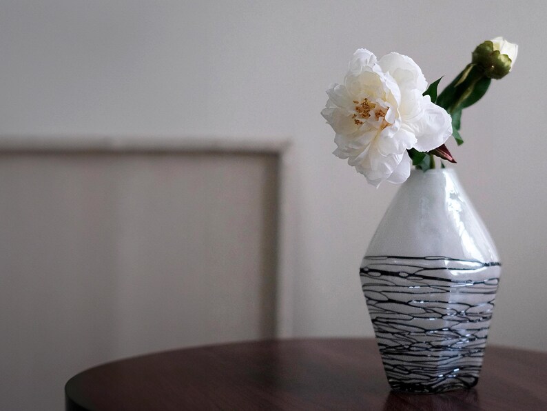 Pode incluir: Um vaso decorativo branco e preto com uma flor de pe&oacute;nia branca e um bot&atilde;o floral. O vaso tem um acabamento brilhante com um design linear preto na metade inferior. O vaso est&aacute; sobre uma mesa de madeira castanha escura.