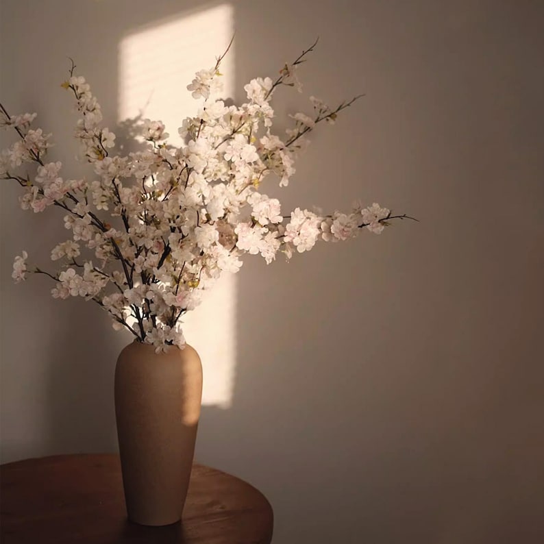 May include: A beige ceramic vase holds a bouquet of white and light pink cherry blossom branches. The vase sits on a wooden table, with sunlight casting shadows on the wall behind it. The overall aesthetic is soft and natural.