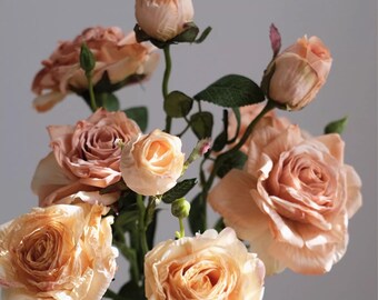 May include: A bouquet of peach-colored roses in various stages of bloom, from tight buds to fully opened flowers. The roses have multiple layers of delicate petals, with green stems and leaves. The overall arrangement is soft and romantic.