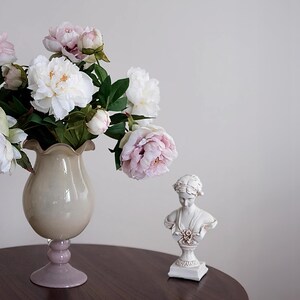 Pode incluir: Uma natureza morta com um vaso de pe&oacute;nias rosa e brancas, um busto decorativo e uma mesa castanha escura. O vaso tem um corpo bege claro e uma base lavanda. O busto &eacute; branco com detalhes dourados.