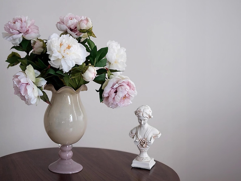 Pode incluir: Uma natureza morta com um vaso de pe&oacute;nias rosa e brancas, um busto decorativo e uma mesa castanha escura. O vaso tem um corpo bege claro e uma base lavanda. O busto &eacute; branco com detalhes dourados.