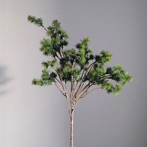 May include: A single artificial evergreen stem with multiple branches. The stem is a light brown color, and the branches are covered in small, spiky green leaves. The background is a solid, neutral gray.