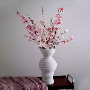 May include: A white, textured vase holds a bouquet of pink and white cherry blossom branches. The vase has a unique, swirling design. A mauve fabric is draped on a dark surface in the foreground. The arrangement is set against a neutral background.