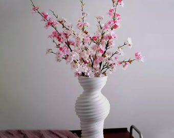 May include: A white, textured vase holds a bouquet of pink and white cherry blossom branches. The vase has a unique, swirling design. A mauve fabric is draped on a dark surface in the foreground. The arrangement is set against a neutral background.