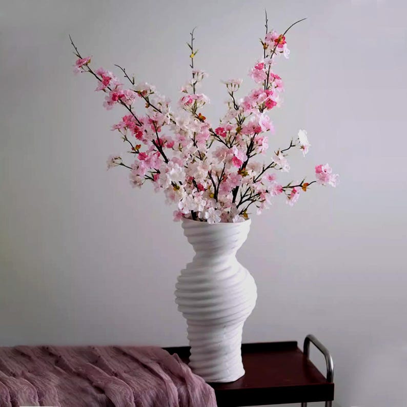 May include: A white, textured vase holds a bouquet of pink and white cherry blossom branches. The vase has a unique, swirling design. A mauve fabric is draped on a dark surface in the foreground. The arrangement is set against a neutral background.