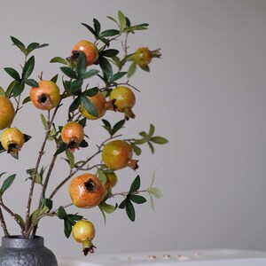 May include: A decorative floral arrangement featuring branches with green leaves and several pomegranates in varying stages of ripeness. The pomegranates are a mix of yellow and orange hues, and the arrangement is in a dark gray vase.