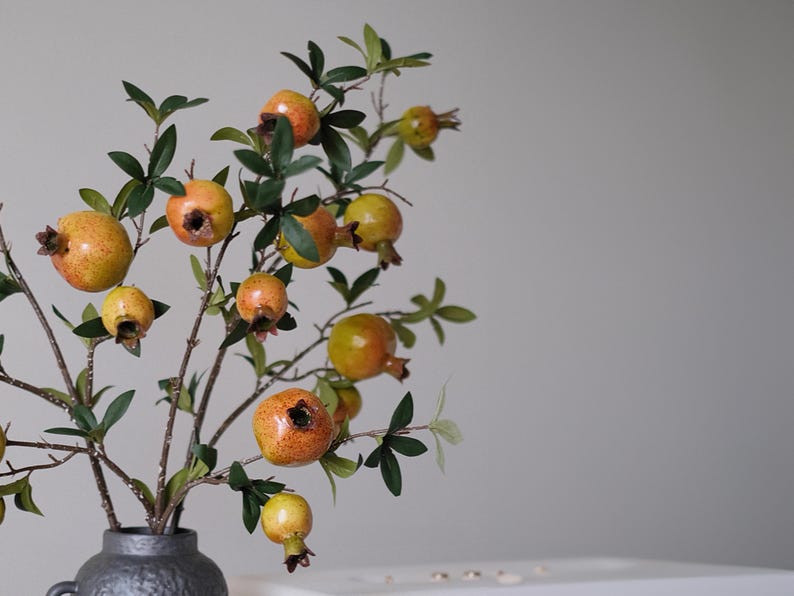 May include: A decorative floral arrangement featuring branches with green leaves and several pomegranates in varying stages of ripeness. The pomegranates are a mix of yellow and orange hues, and the arrangement is in a dark gray vase.